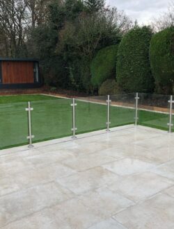 Porcelain tiled patio with glass balustrade overlooking turfed garden and garden room